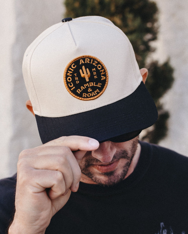 Man tipping the cream and black The Shop Two Tone Hat with a round Iconic Arizona Ramble and Roam patch