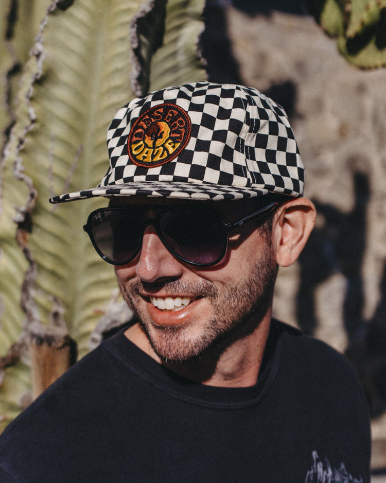 Smiling man in sunglasses wearing the black and white Desert Daze Checkerboard Hat with a round skull patch