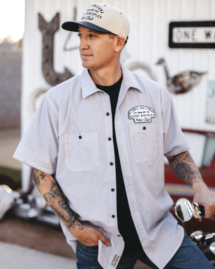 Man in the striped Moto Club Mechanic Shirt with Desert Disciples chest patch standing by a motorcycle