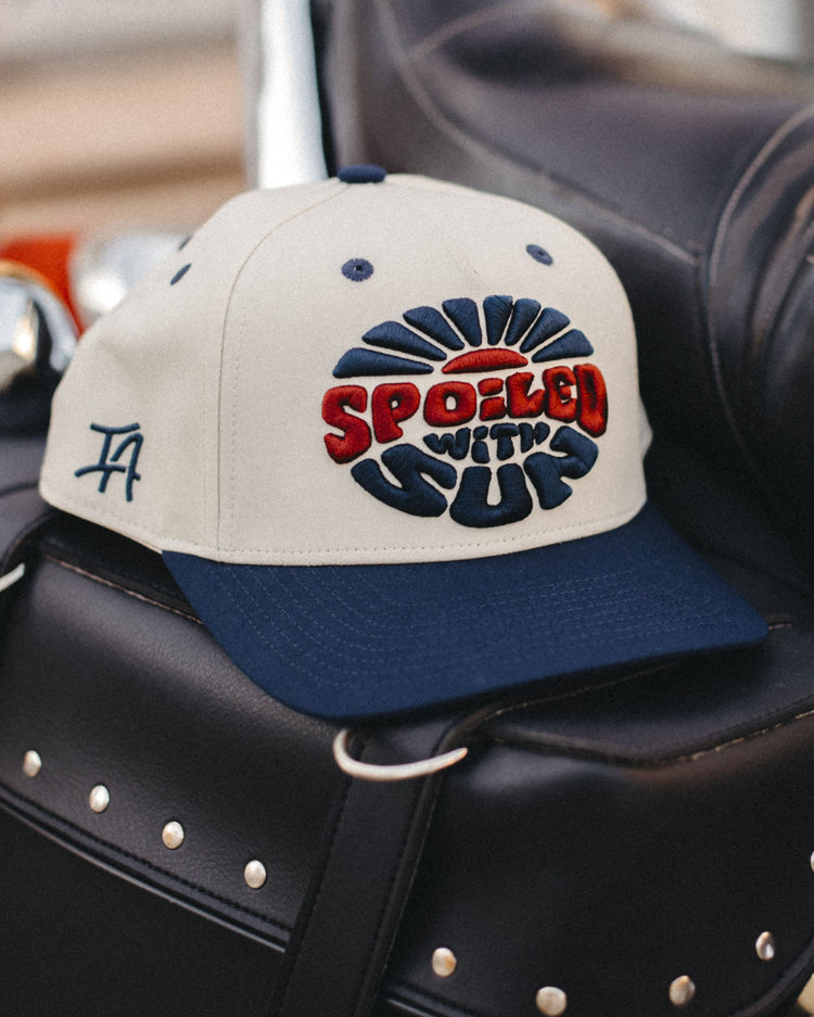 Cream and navy Spoiled With Sun Two Tone Hat with red and navy sun design on a leather motorcycle seat