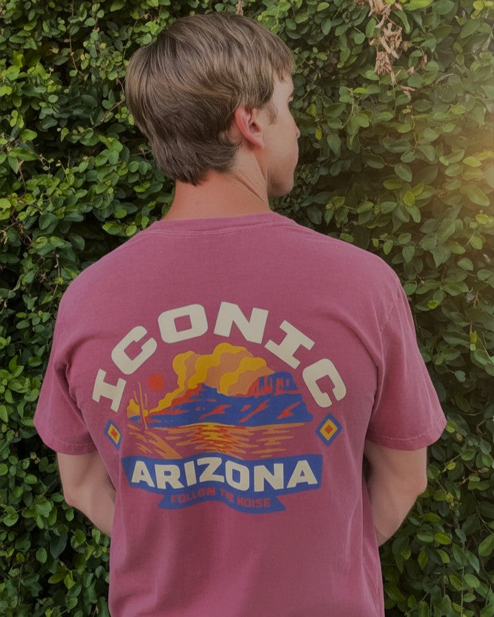 A man viewed from behind wearing a dusty rose t-shirt with a large graphic on the back that reads 'ICONIC ARIZONA' with a desert landscape illustration, set against a background of green foliage.